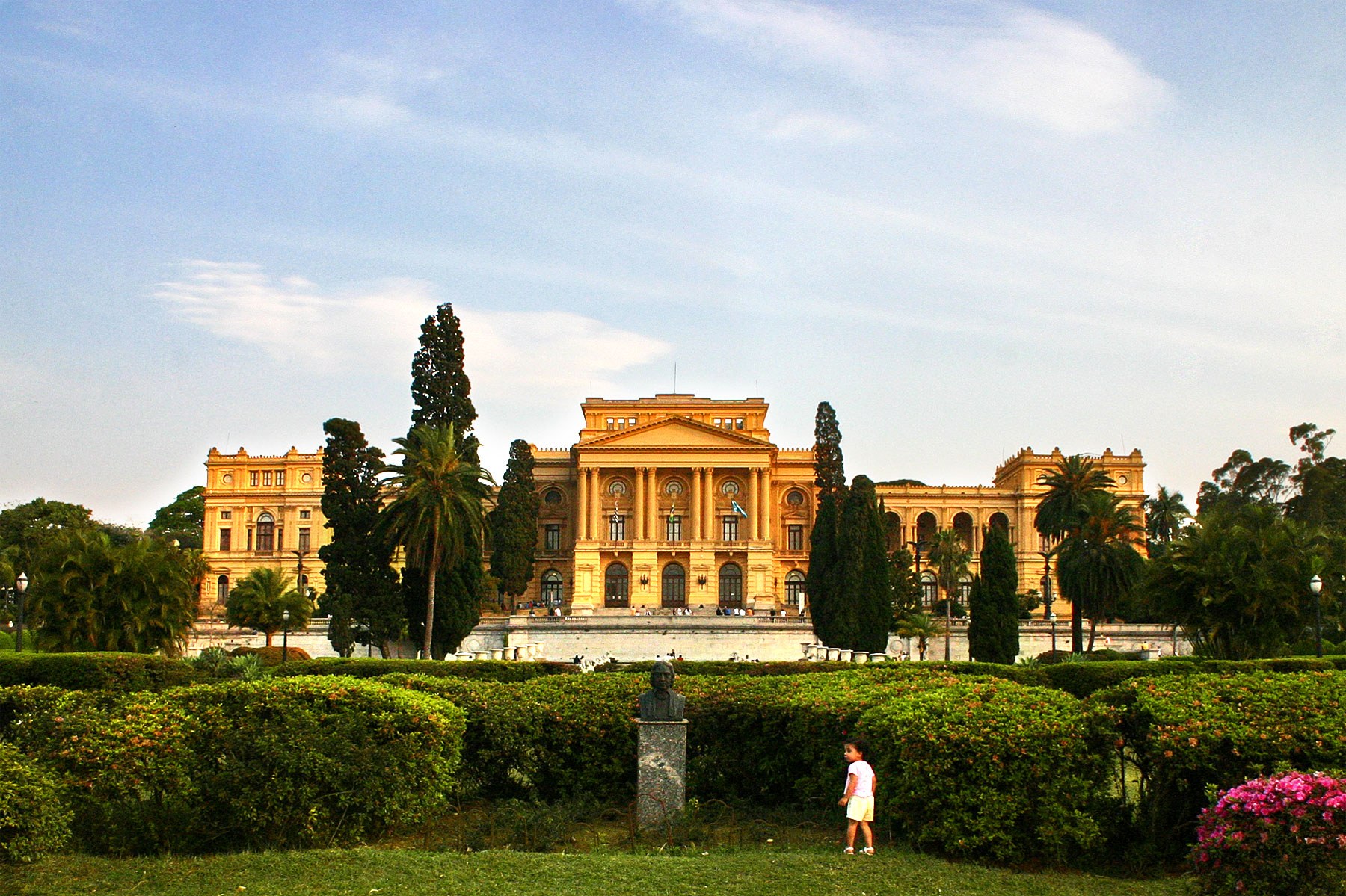 Fachada amarela do Museu do Ipiranga com jardim, palmeiras e ciprestes, em São Paulo.